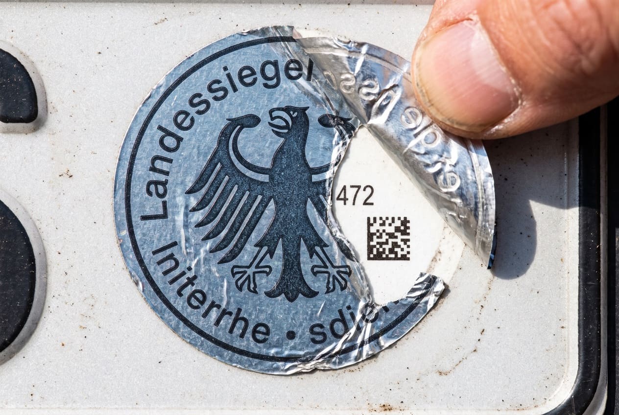 Teilweise abgezogene Stempelplakette auf dem Kennzeichen mit sichtbarem 3-stelligem Code